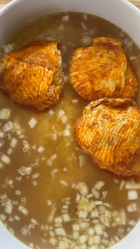 Three seasoned chicken thighs with onions in a broth-filled white bowl.