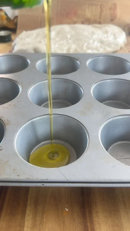 Pouring olive oil into a muffin tin cup, with dough wrapped in plastic on the table.