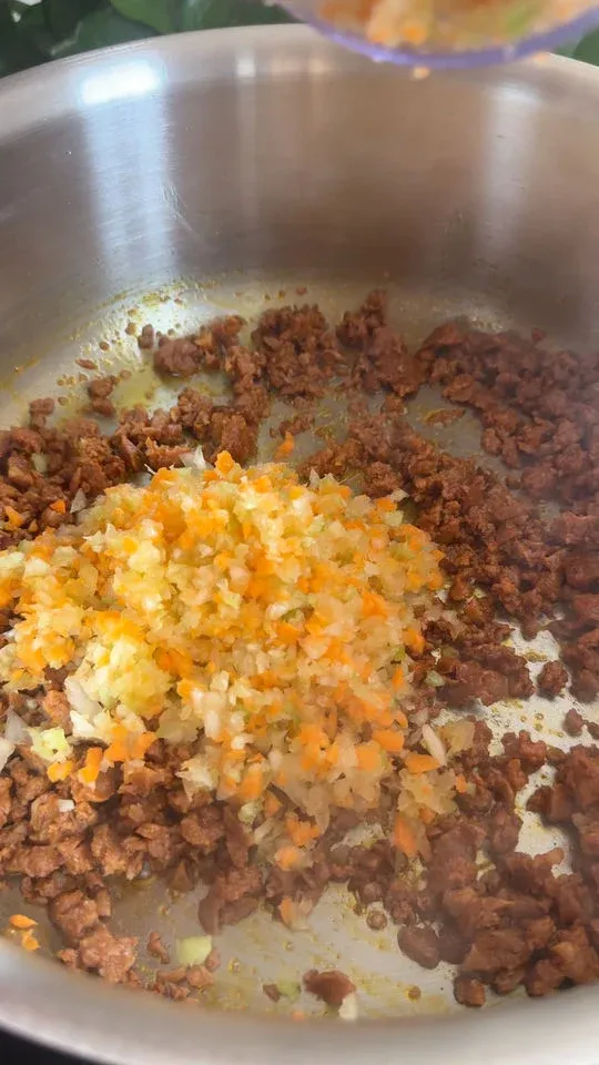 Chopped onions and carrots sautéing with ground meat in a stainless steel pan.