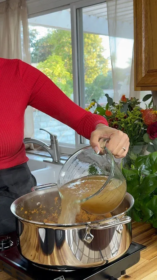 Person in red shirt pouring broth into a stainless steel pot, with a kitchen sink and window in the background.