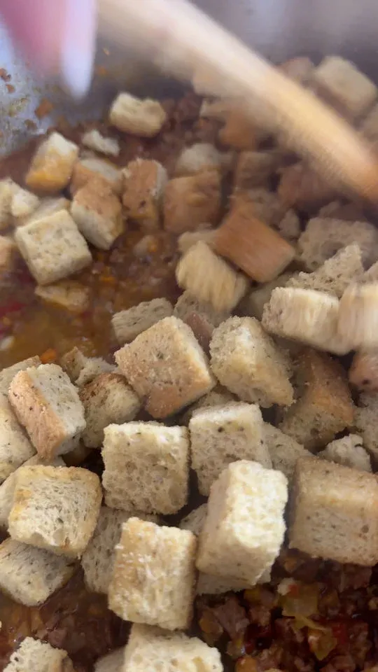 Stuffing being stirred into a mixture of cooked vegetables and meat in a pot.