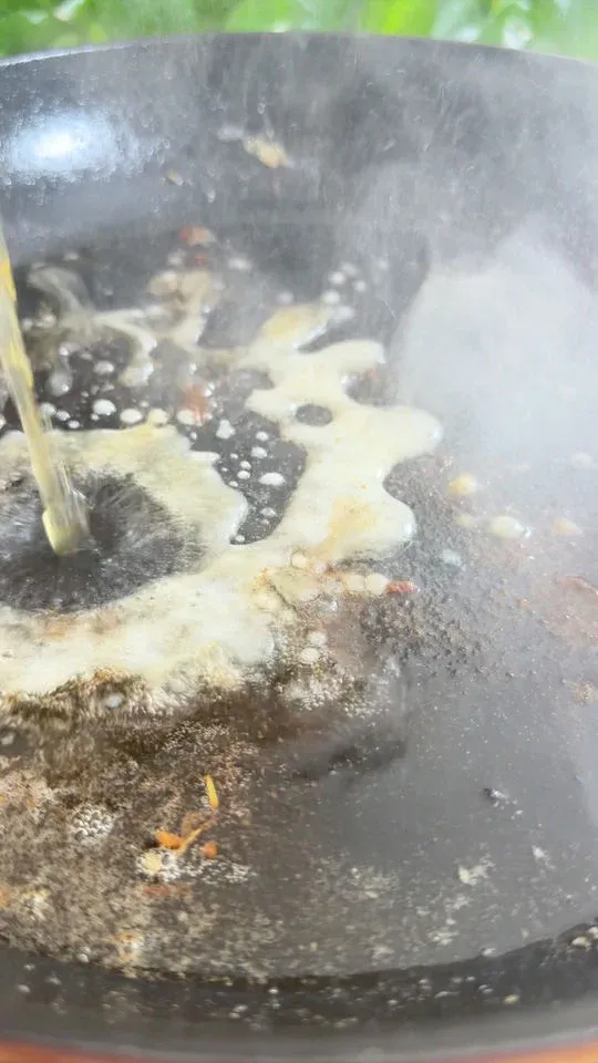 Pouring liquid into a steaming hot pan with sizzling oil and spices.