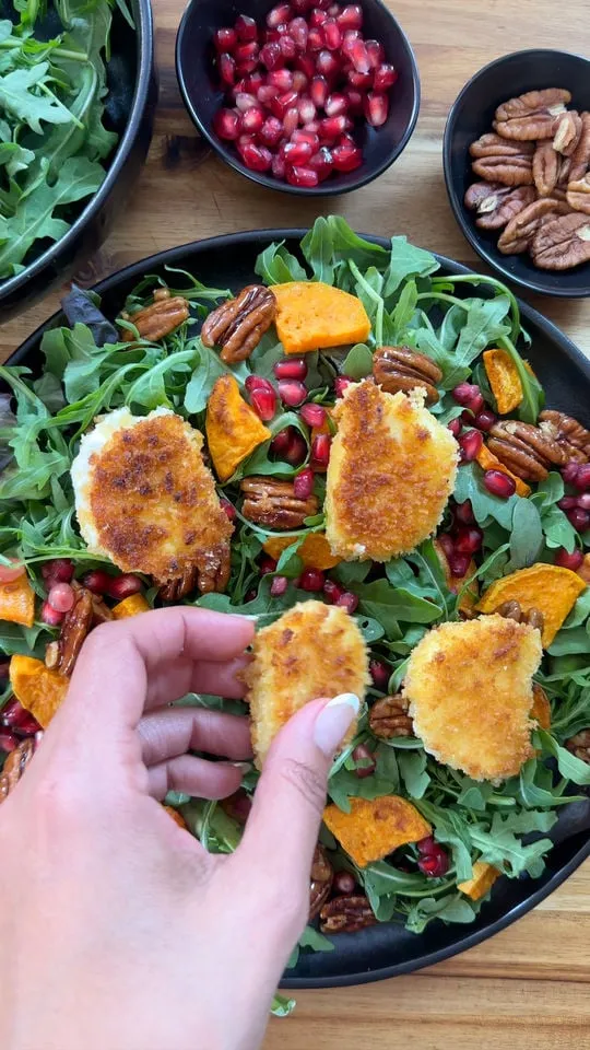 Hand picking fried goat cheese from salad with arugula, pecans, pomegranate, and sweet potato, bowls in background.