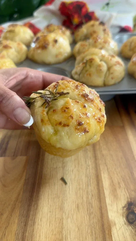 Hand holding a freshly baked focaccia muffin topped with rosemary on a wooden table background.