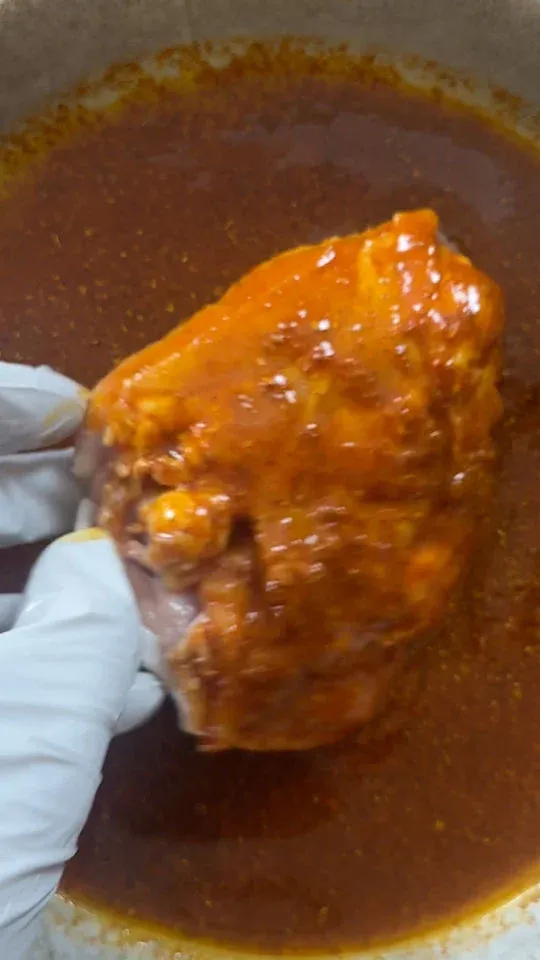 Gloved hand holding marinated chicken thigh over bowl with marinade.