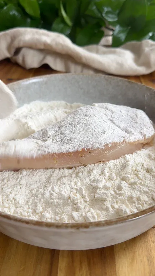 Raw chicken breast being coated with flour in a shallow bowl on a wooden surface.