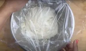 Bowl with dough covered in plastic wrap on a wooden surface, hands holding the edge of the bowl.