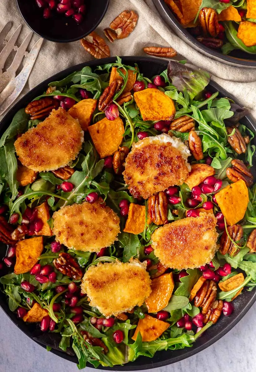 Crispy goat cheese salad with fried goat cheese, arugula, sweet potatoes, pecans, and pomegranate seeds in a black bowl.