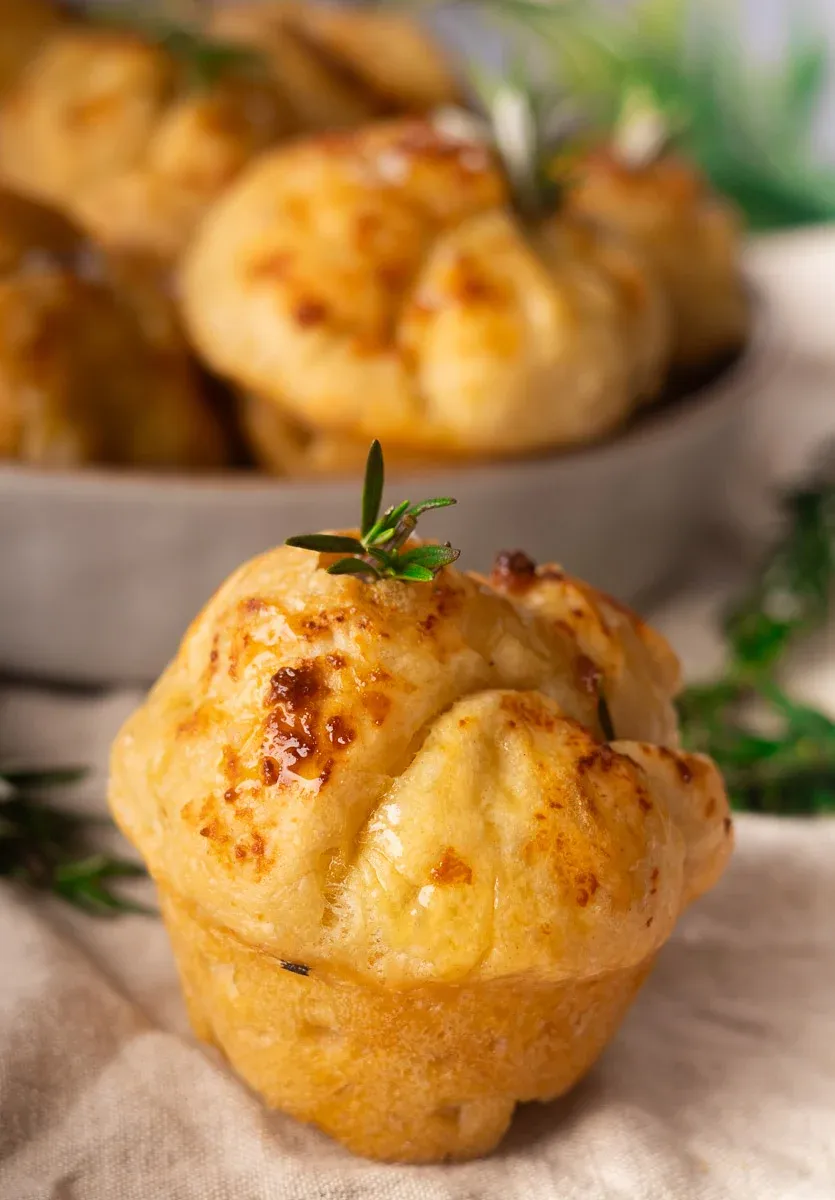 Golden-brown No-Knead Focaccia muffins garnished with rosemary, set on a beige cloth, with more muffins in the background.