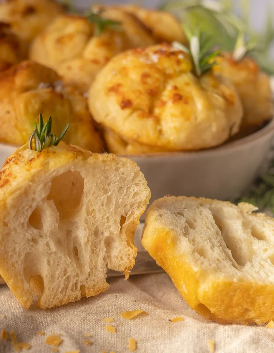 Close-up of freshly baked No-Knead Focaccia muffins with rosemary, one split open to show texture, placed on a cloth.