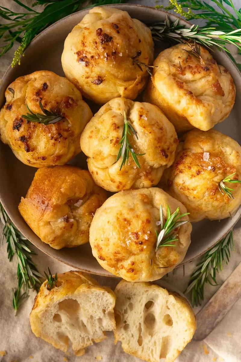 No-Knead Focaccia muffins with rosemary on a plate, surrounded by fresh rosemary sprigs.