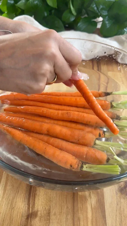 Hand washing fresh carrots in a glass bowl filled with water on a wooden surface.