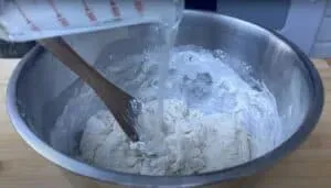 Pouring water into a mixing bowl of flour with a wooden spoon.