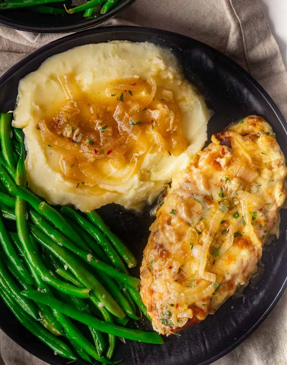 French onion soup chicken with caramelized onions, mashed potatoes, and green beans on a black plate.
