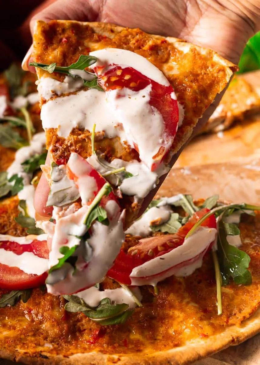Hand holding slice of Lebanese Chicken Pizza with tomato, arugula, and creamy sauce on a wooden board.
