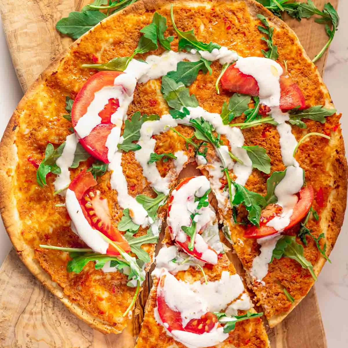 Lebanese chicken pizza with tomato slices, arugula, and white sauce drizzle on a wooden board.