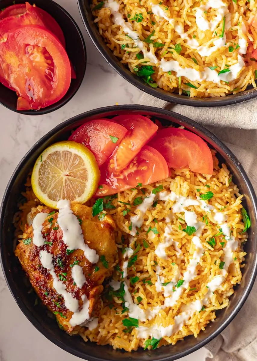 Mediterranean Baked Chicken Thighs and Rice with tomatoes, lemon, and sauce in black bowls on marble surface.
