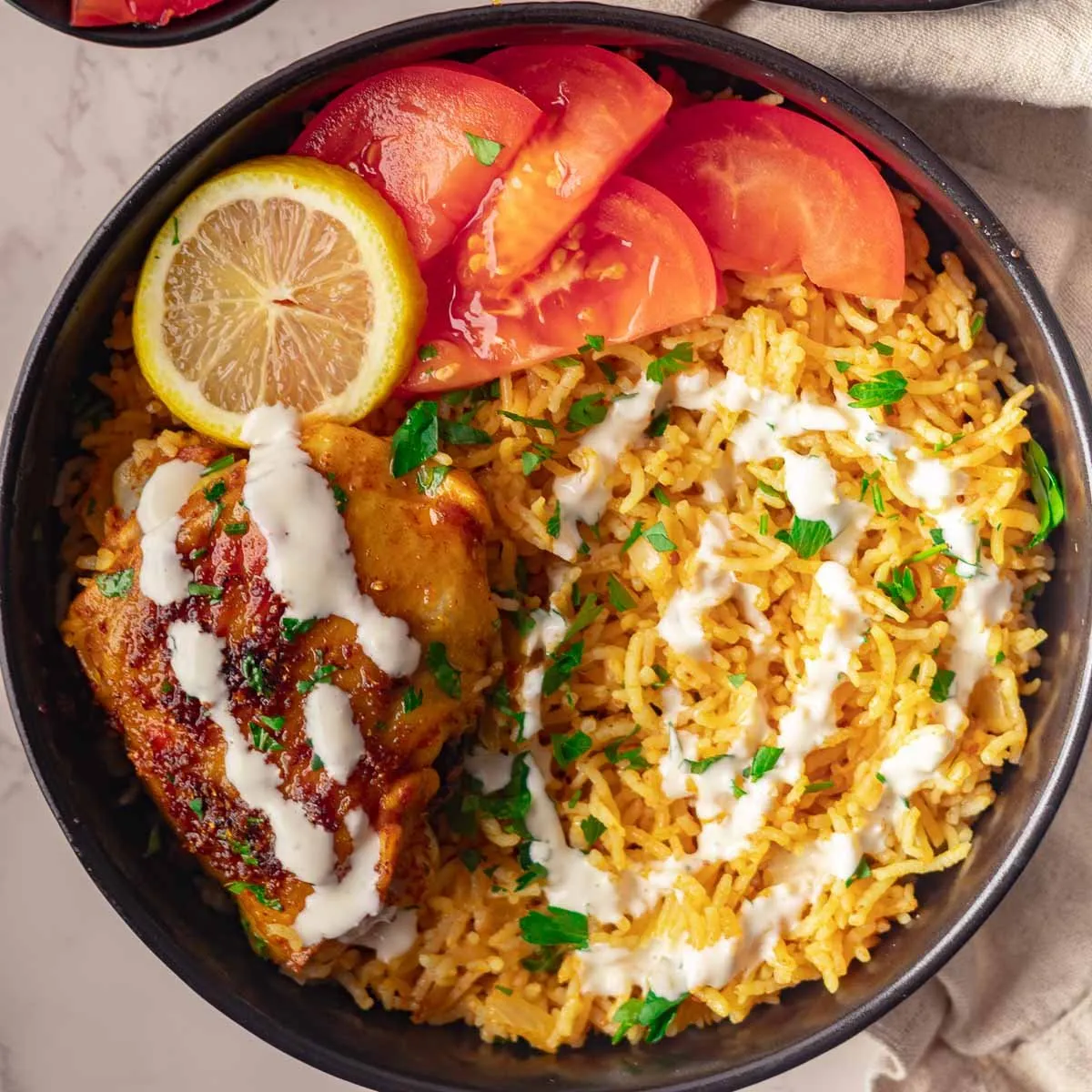 Grilled chicken with yogurt sauce, yellow rice, tomato slices, and a lemon wedge in a black bowl.