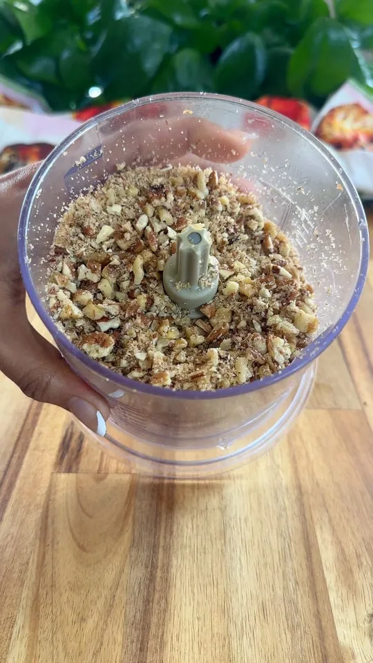 Crushed walnuts in a food processor held by a hand over a wooden table.