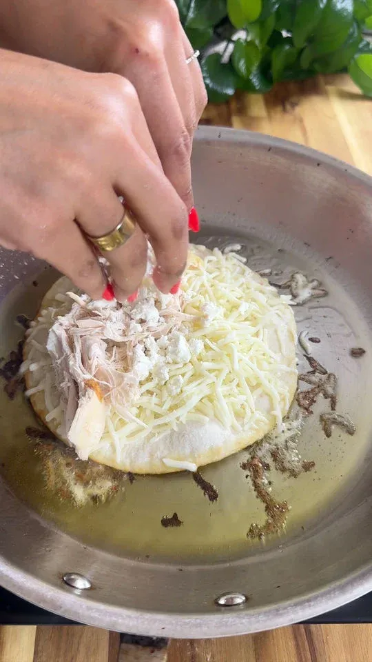 Hands adding shredded chicken and cheese to a tortilla in a frying pan.