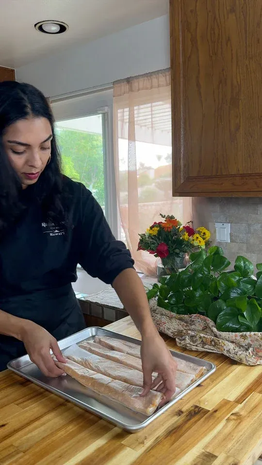 Woman arranging meat logs on a baking tray in a kitchen, with flowers and a plant in the background.