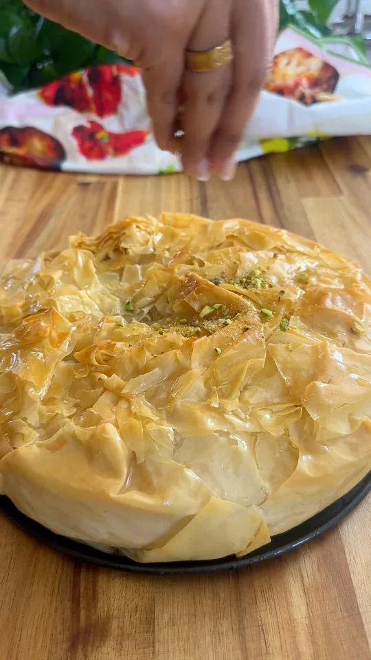 Hand sprinkling pistachios on a golden, flaky baklava pie on a black plate, wooden table underneath.