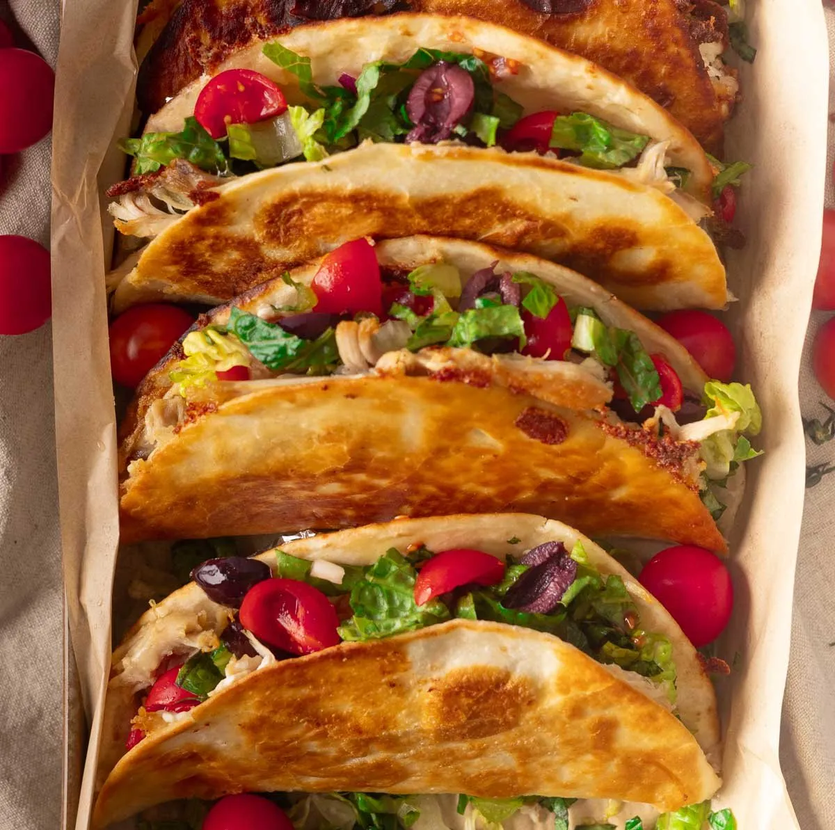 Greek crispy chicken tacos filled with lettuce, tomatoes, olives, and shredded chicken on a parchment-lined tray.