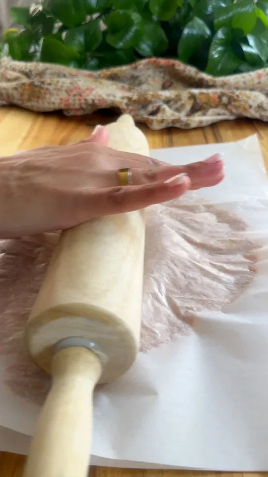 Hand using a rolling pin on parchment paper with greenery in the background.