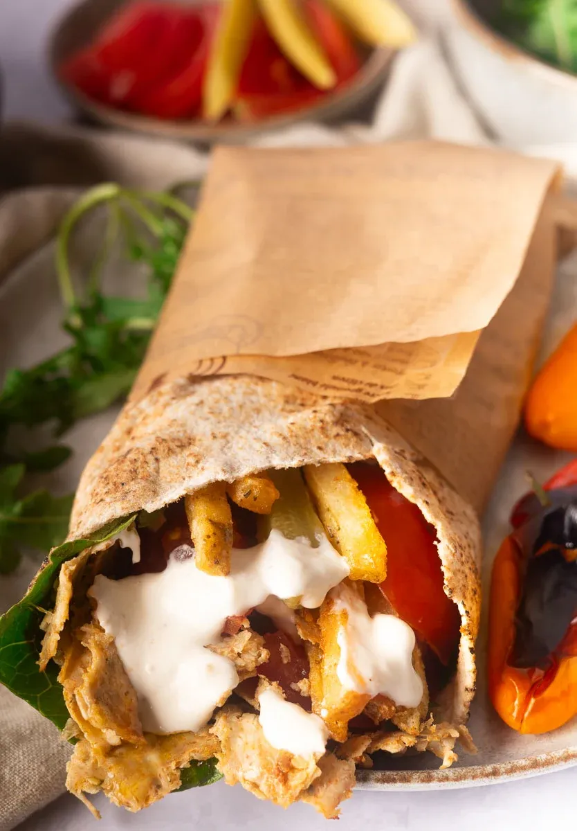Close-up of a wrapped Viral doner kebab with fried potatoes, sauce, and vegetables next to roasted peppers on a plate.