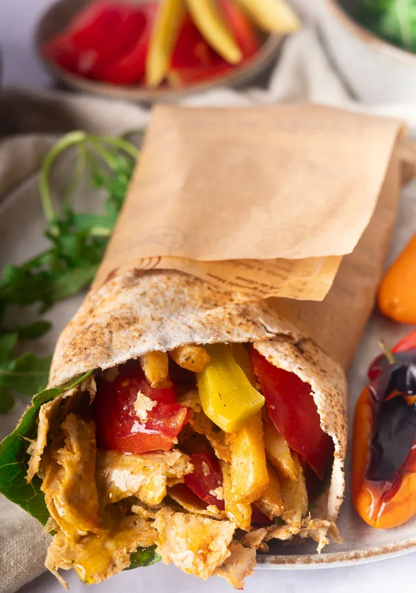 Viral doner kebab wrapped in pita in brown paper with grilled chicken, peppers, and fries on a plate with herbs.