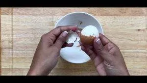Hands peeling a boiled egg over a bowl on a wooden table.