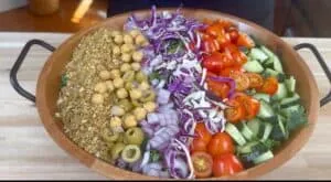 Colorful salad bowl with cherry tomatoes, cucumbers, onions, olives, chickpeas, cabbage, and breadcrumbs on a wooden table.