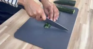 Person slicing cucumber on a gray cutting board, wooden kitchen counter background.