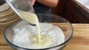 Pouring batter into a glass bowl of flour on wooden countertop, ready for baking.