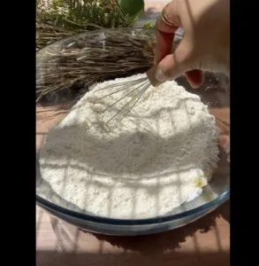 Hand whisking flour in a glass bowl with natural light and wheat decor.