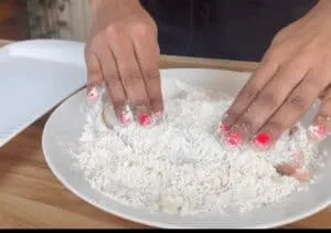 Hands coating chicken in flour on a white plate, preparing for frying in a kitchen setting.