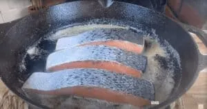 Three salmon fillets frying in a cast iron skillet.