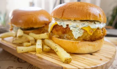 Golden crispy cod fish sandwich with cheese and dill sauce, served with seasoned fries on a wooden platter.