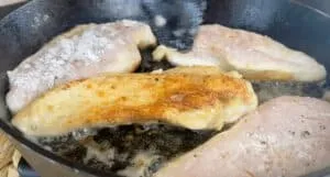 Chicken breasts frying in a skillet, partially browned and seasoned, close-up shot.