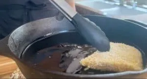 Tongs holding breaded food over a hot skillet in the kitchen, demonstrating frying techniques.