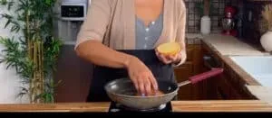 Person cooking with a frying pan, placing a bun in the pan in a cozy kitchen setting.