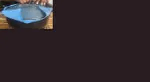 Person removing a glass lid with a blue cloth from a steaming black pot.