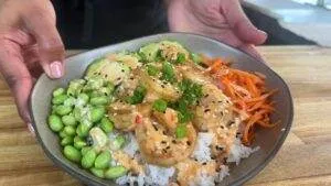 Colorful shrimp poke bowl with rice, edamame, carrots, cucumber, and creamy sauce, garnished with green onions.