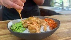 Close-up of a shrimp rice bowl with edamame, carrots, and spicy mayo being drizzled on top on a wooden table.
