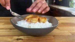 Person plating grilled shrimp on white rice in a bowl, showcasing a delicious homemade seafood meal preparation.
