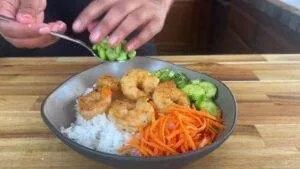A delicious shrimp rice bowl with carrots, cucumbers, and edamame being served on a wooden table.