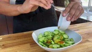 Person adding dressing to fresh cucumber salad in a white bowl on a wooden countertop.