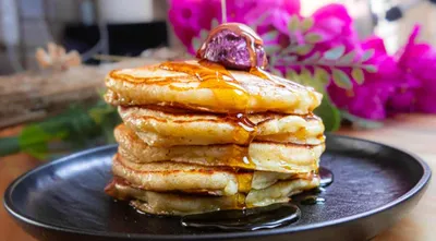 Stack of fluffy pancakes drizzled with syrup, topped with a berry, and set against a floral background.