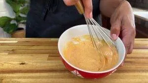 Whisking creamy orange sauce in a red bowl on a wooden countertop.