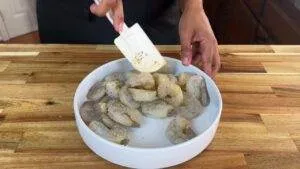 Person seasoning shrimp in a white bowl with a spatula on a wooden kitchen counter.